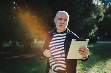 Sıradan giyinmiş, elinde gözlükle parkta durup tablet üzerinde internete göz gezdirip kameraya bakarken konsantre olmuş, ciddi bir erkek.