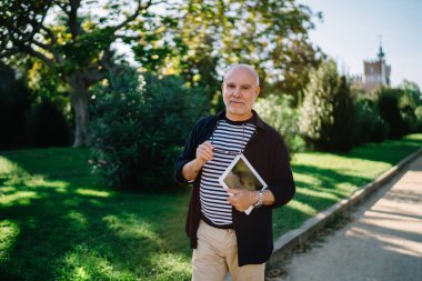 Gün ışığında parkta yürüyüp kameraya bakarken elinde tablet ve gözlüklerle gülümseyen yaşlı bir erkek.