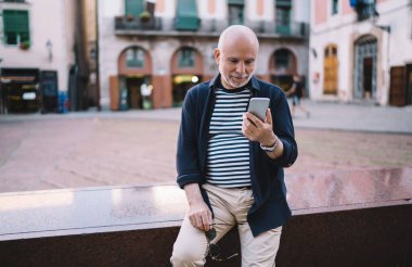 Sınıra yaslanırken ve şehir merkezinde cep telefonuna bakarken günlük giysiler içinde gülümseyen yaşlı adam güneş gözlüğü takıyor.