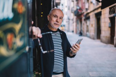 Gün ışığında şehir caddesinde durup kameraya bakarken elinde gözlük olan ve akıllı telefon kullanan konsantre olmuş kel bir adam.
