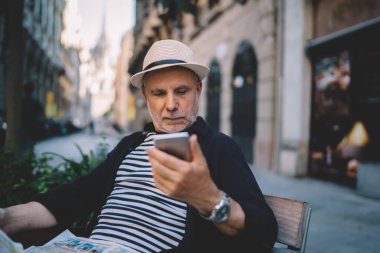 Rahat giyinen ve şapka takan olgun bir adam akıllı telefondan mesajları kontrol ediyor sokak kafesinin masasında oturmuş gazete okuyor.