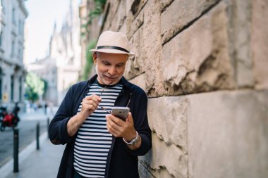 Gülümseyen olgun erkek, tuğla duvarın yanındaki kaldırımda dururken internet üzerinden bilgi okuyan cep telefonu ekranına odaklanmış.
