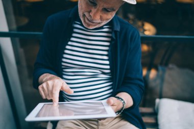 Gri sakallı üst düzey bir adam gündelik giysiler içinde modern kafenin penceresinin yanındaki bankta oturuyor ve tablet kullanırken bilgileri kontrol ediyor.