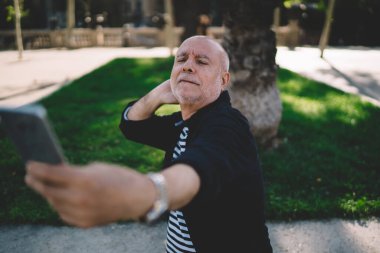 Gri sakallı, neşeli, yaşlı bir adam parkta yürürken ayakta durup cep telefonu kullanıyor.