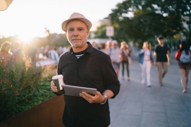 50 yaşında erkek, günlük kıyafet giymiş elinde bir fincan kahve ve tabletle şehrin yeşil bitkilerinin yanında duruyor.