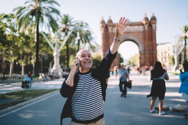 Sıradan kıyafetlerle gülümseyen orta yaşlı bir erkek cep telefonuyla konuşuyor ve sokakta dururken el ele el sallıyor.