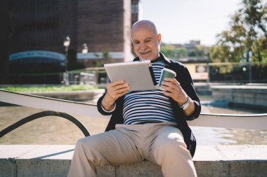 Moda kıyafetleri içinde gülümseyen yaşlı bir adam göletin yanında tek kullanımlık kahve bardağıyla bankta oturuyor ve caddede tablet tarıyor.