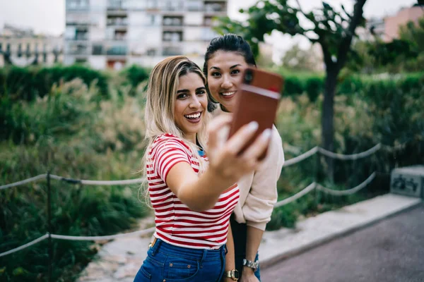 笑着向高加索和亚洲嬉皮士女孩们摆出一副智能手机相机的架势 利用空闲时间为博客制作内容 快乐的20多岁的年轻女性在汤姆的空闲时间里通过手机应用程序自拍 — 图库照片