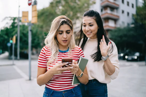 Multiracial hipster girls enjoying mobility chatting and messaging during city sightseeing on vacations, charming female bloggers using smartphone for making online booking and travel ticketing