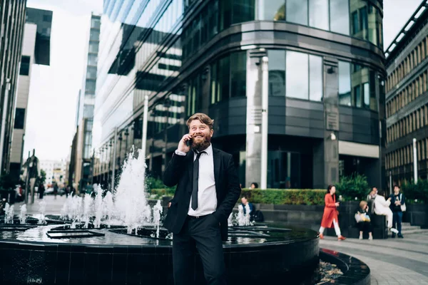 身穿正装 打着领带 头戴大胡子的商人站在现代建筑附近的街上望着远方 一边用手机交谈 — 图库照片