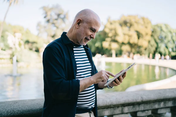 Sıradan giyinmiş, taş tırabzanların yanında duran ve parkta yürürken tablet üzerinde sohbet eden yaşlı adamın yan görüntüsü.