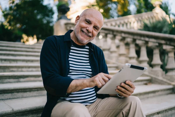 穿着休闲装的快乐老人 一边对着相机微笑 一边坐在公园的石阶上 一边在平板电脑上冲浪 — 图库照片