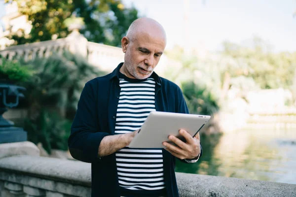Gri sakallı ve kel kafalı ciddi yaşlı bir adam şehir parkında dinlenirken taş parmaklıkların yanında durup tablet karıştırıyor.
