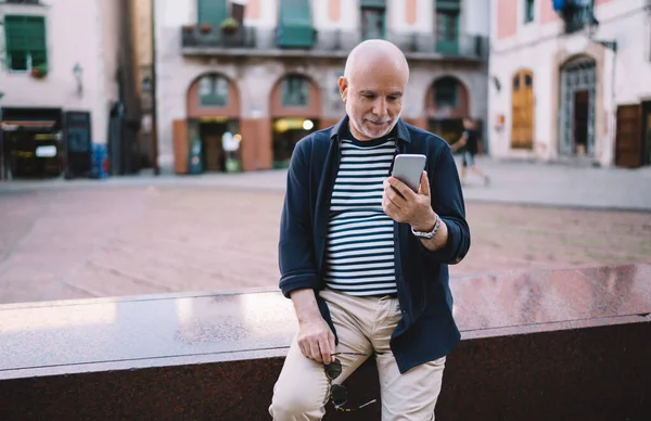 Sınıra yaslanırken ve şehir merkezinde cep telefonuna bakarken günlük giysiler içinde gülümseyen yaşlı adam güneş gözlüğü takıyor.