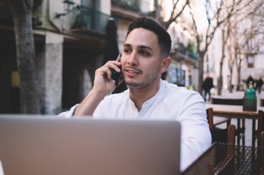 Resmi olarak kendine güvenen etnik erkek serbest çalışan, açık kafedeki ahşap masada oturan ve iş sırasında laptopla akıllı telefondan arayan biri.