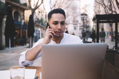 Açık kafedeki ahşap masada oturan ve proje üzerinde çalışırken telefonla konuşan kendine güvenen serbest çalışan etnik bir erkek.