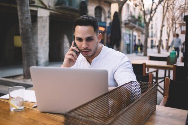Resmi olarak odaklanmış etnik erkek serbest giyiniyor açık kafedeki ahşap masada oturuyor ve dizüstü bilgisayar projesinde çalışırken akıllı telefondan arıyor.