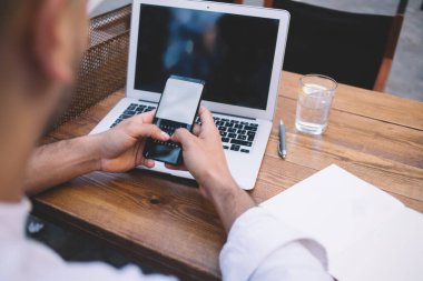 Yüksek açılı beyaz gömlekli serbest çalışan, açık kafede dizüstü bilgisayarlı, tahta masada oturan ve akıllı telefondan boş ekranlı mesajlaşan bir erkek.