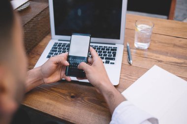 Tahta masada dizüstü bilgisayarla oturan ve açık kafedeki akıllı telefondan mesajlaşan yüksek açılı isimsiz serbest çalışan bir erkek.