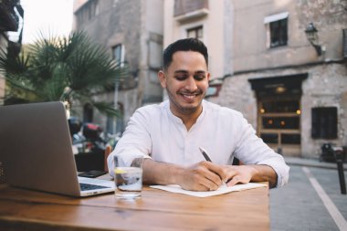 Havalı kıyafetli iyimser bir adam rahat bir terasta oturuyor ve öğle yemeği molasında kalemle kağıt üzerine rapor yazıyor.