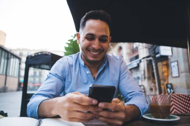 Rahat bir kafede çay içerken gülümseyen, mesajlaşan, gündelik kıyafetli, pozitif, genç bir adam.