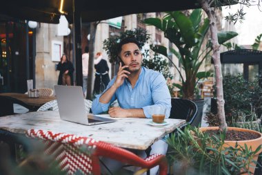 Düşünceli, serbest çalışan etnik bir erkek, ahşap masada dizüstü bilgisayar ve kahveyle otururken telefonla konuşuyor ve gözlerini kaçırıyor.