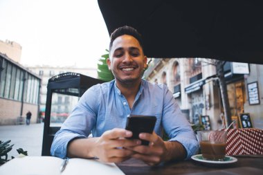 Açık mavi tişörtlü, gülümseyen genç bir öğrenci kaldırım kafesinde içki içerken cep telefonunu karıştırıyor.