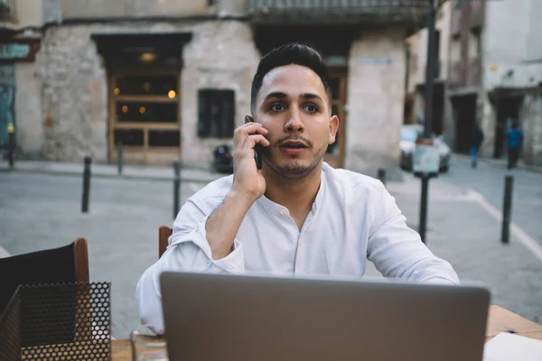 Thoughtful ethnic man speaking on cellphone with interest about new work tasks and sitting in creative workplace while looking away