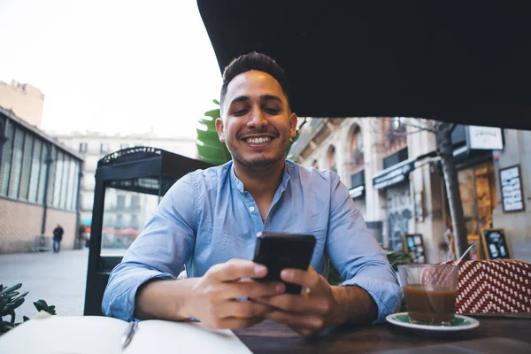 Młody Uśmiechnięty Student Etniczny Jasnoniebieskiej Koszuli Przeglądający Telefon Komórkowy Podczas — Zdjęcie stockowe