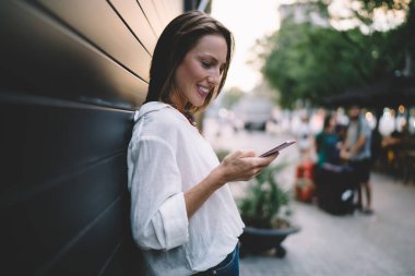 Modayı takip eden yetişkin kadınların yan görünüşü, kentteki modern binaların duvarına yaslanırken ve ayakta dururken cep telefonlarında mesajlaşma takınıyor