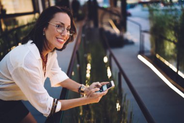 Şehir caddesinde tırabzanlara yaslanmış şık giyinmiş, sosyal ağdaki haberleri akıllı telefondan kontrol eden pozitif kadın.