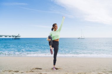 40 yaşında esmer bir kadın. Kumlu deniz kenarında egzersiz yaparken dizleri göğsünde, kolları havada duruyor.