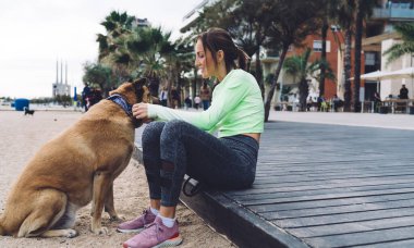 Yürüyüş sırasında ahşap patikada dinlenirken köpek tasması tutan spor elbiseli, pozitif genç bir kadının yan görüntüsü.
