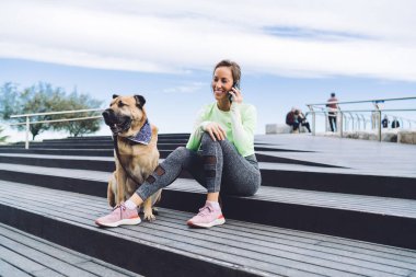 Modayı takip eden spor kıyafetleri içinde mutlu bir bayan antrenör sabah vakti akıllı telefon aracılığıyla uluslararası sohbetin tadını çıkarıyor. Saf köpeği gezdiren neşeli ve sağlıklı bir kız.