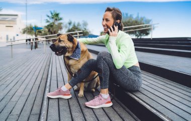 Modayı takip eden spor kıyafetleri içinde neşeli bir bayan antrenör sabah vakti akıllı telefon aracılığıyla uluslararası sohbetin tadını çıkarıyor. Safkan köpeği gezdiren neşeli ve sağlıklı bir kız.