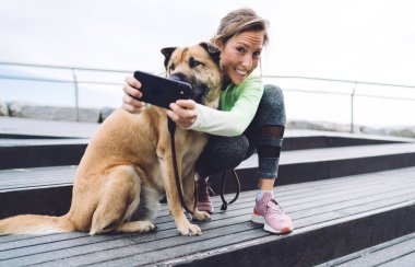 Aktif giyimli neşeli olgun bir kadının merdivenlerde köpekle oturması ve kameraya bakarken cep telefonunu karıştırması.