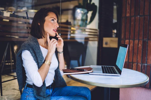 Uzaktan kumanda sırasında kafede oturan, bilgisayar kullanan, ciddi beyaz kadın, akıllı telefon konuşmalarıyla rezervasyon ve teslimatı onaylayan düşünceli kadın.