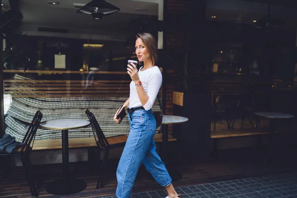 街のカフェを背景に街歩きやコーヒーを飲みながらノートを手に白いトップと青いジーンズの美しい女性 — ストック写真