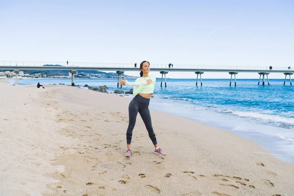 海岸線でのウォーミングアップ中に体の筋肉を伸ばす朝のジョギングのためのトラックスーツの強い女性トレーナー 穏やかな海岸でのレジャー時間の間に屋外のピラティスを楽しむ柔軟なフィットの女の子 — ストック写真