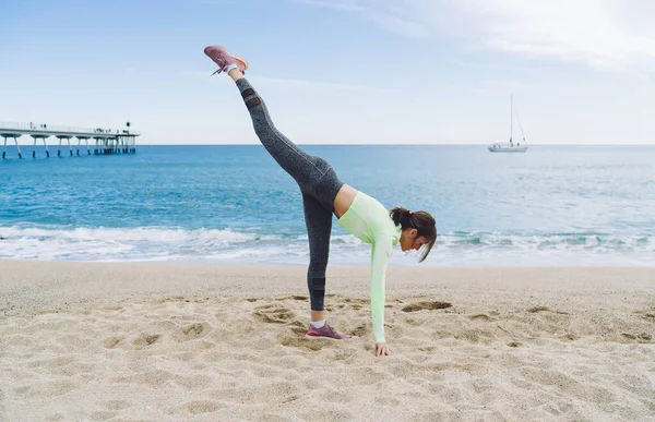 Female slim woman in active wear doing exercise with raising leg and breathing at seashore coastline, young fit girl with flexible and strong body keeping healthy lifestyle with stretching practice