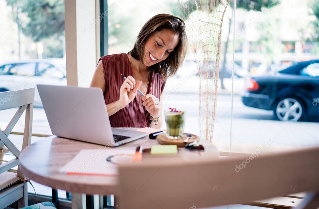 Smiling prosperous female writer in casual wear working remotely in cafe interior making notes in notepad, cheerful prosperous hipster girl creating article for publication in blog using laptop computer