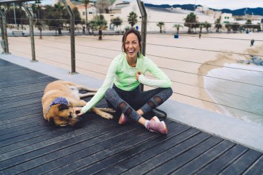 İyimser orta yaşlı kadın spor kıyafetleri içinde. Kereste yolunda otururken sahilde uyuyan köpeğe gülüyor ve onu okşuyor.