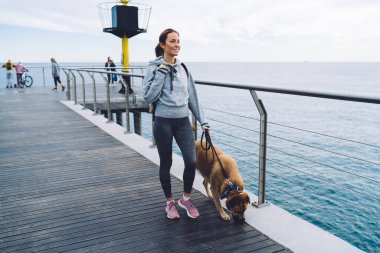 Spor kıyafetli neşeli genç bir kadın tasmalı köpekle yürüyor ve deniz kıyısındaki tatil beldesinde dinlenirken denize bakıyor.