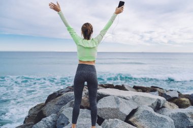 Spor elbiseli genç bir kadının arka planda durup akıllı telefondan müzik dinlemesi ve el kaldırarak deniz dalgalarına hayran olması.
