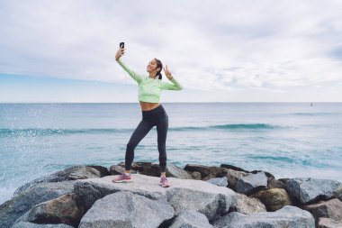 Sahil taşlarında poz veren neşeli kardiyo eğitmeni komik selfie resimleri çekmek için ön mobil kamera kullanıyor, V işaretli kulaklık takan kız video vlog 'u etkiler.