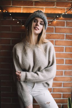 Kış şapkası ve kazak giyen modaya uygun giyinmiş hippi kızın yarım boy portresi boş zamanlarında kameraya bakıyor, kapalı moda fotoğraf çekimi sırasında genç nesil Y poz veriyor.