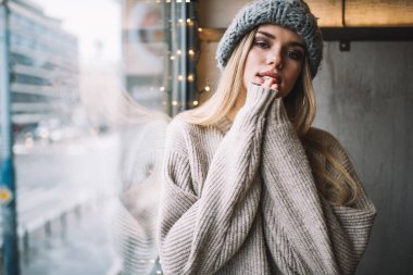 Kış şapkası ve kazak giyen modaya uygun giyinmiş hippi kızın yarım boy portresi boş zamanlarında kameraya bakıyor, moda fotoğraf çekimi sırasında vitrin kenarında genç nesil Y pozu veriyor.