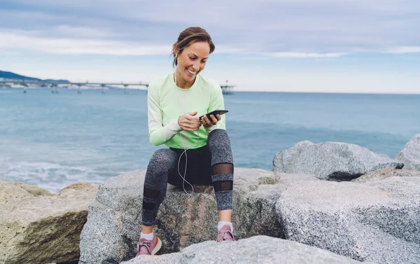 在电子耳机里快乐健康的女孩选择音乐歌曲通过手机应用程序收听 快乐的慢跑者穿着运动服躺在海滨岩石石和连接4G聊天 — 图库照片