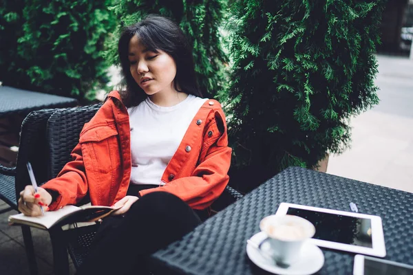 Content young trendy Asian female writing down thoughts in notebook comfortably placing on chair while enjoying fresh cappuccino in street cafe