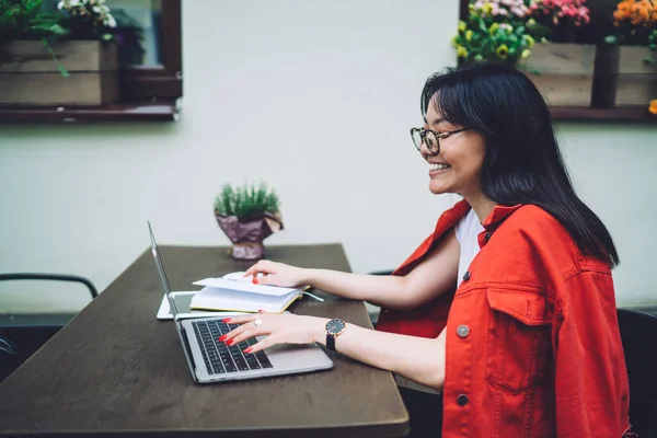 身穿休闲装和眼镜坐在木制桌旁 在笔记本电脑上工作的积极的亚洲女自由职业者的侧视图 — 图库照片